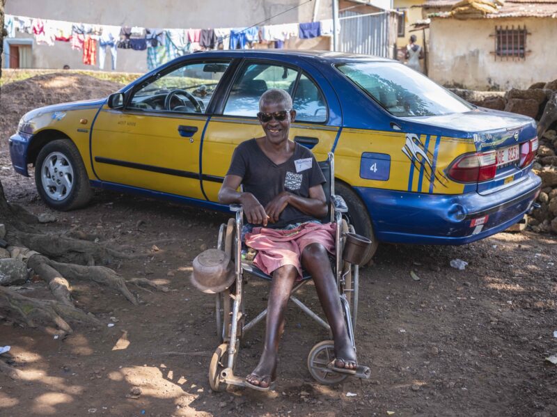 Asmana stting in a wheelchair, smiling . He is in front of a taxi. Cover Image