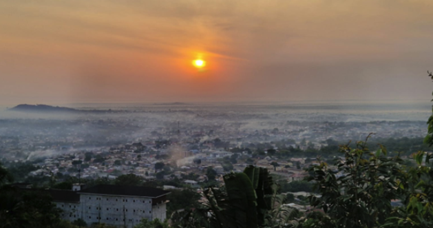 Photo of a glowing orange sun rising over Ho. The foreground shows dense green foliage, while the background is a sprawling mist-covered town extending toward distant hills. Cover Image
