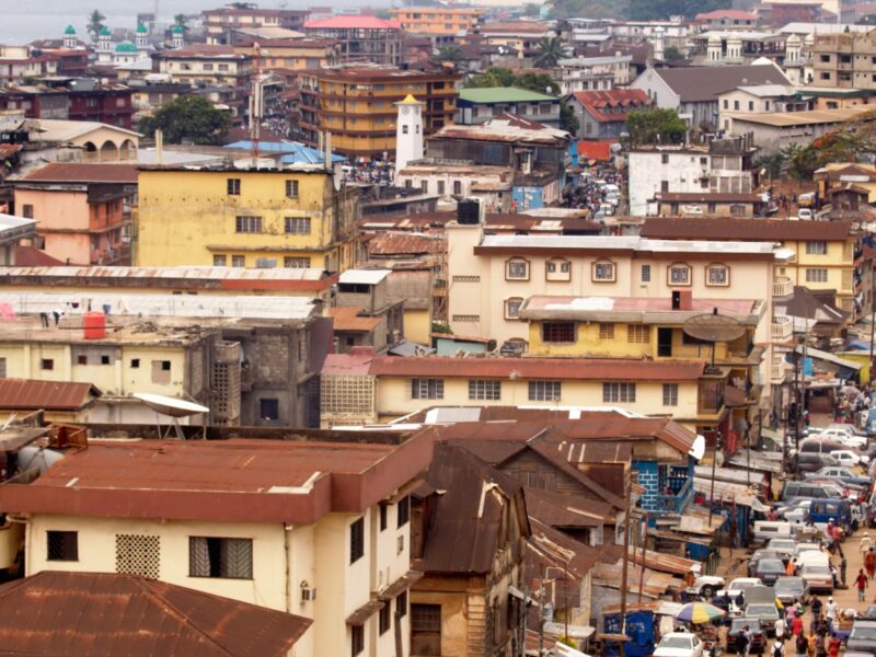 Image of Freetown in Sierra Leone, photo overlooking a section of the city, showing a fairly high concentration of buildings, and a busy roads with traffic and people walking. Cover Image