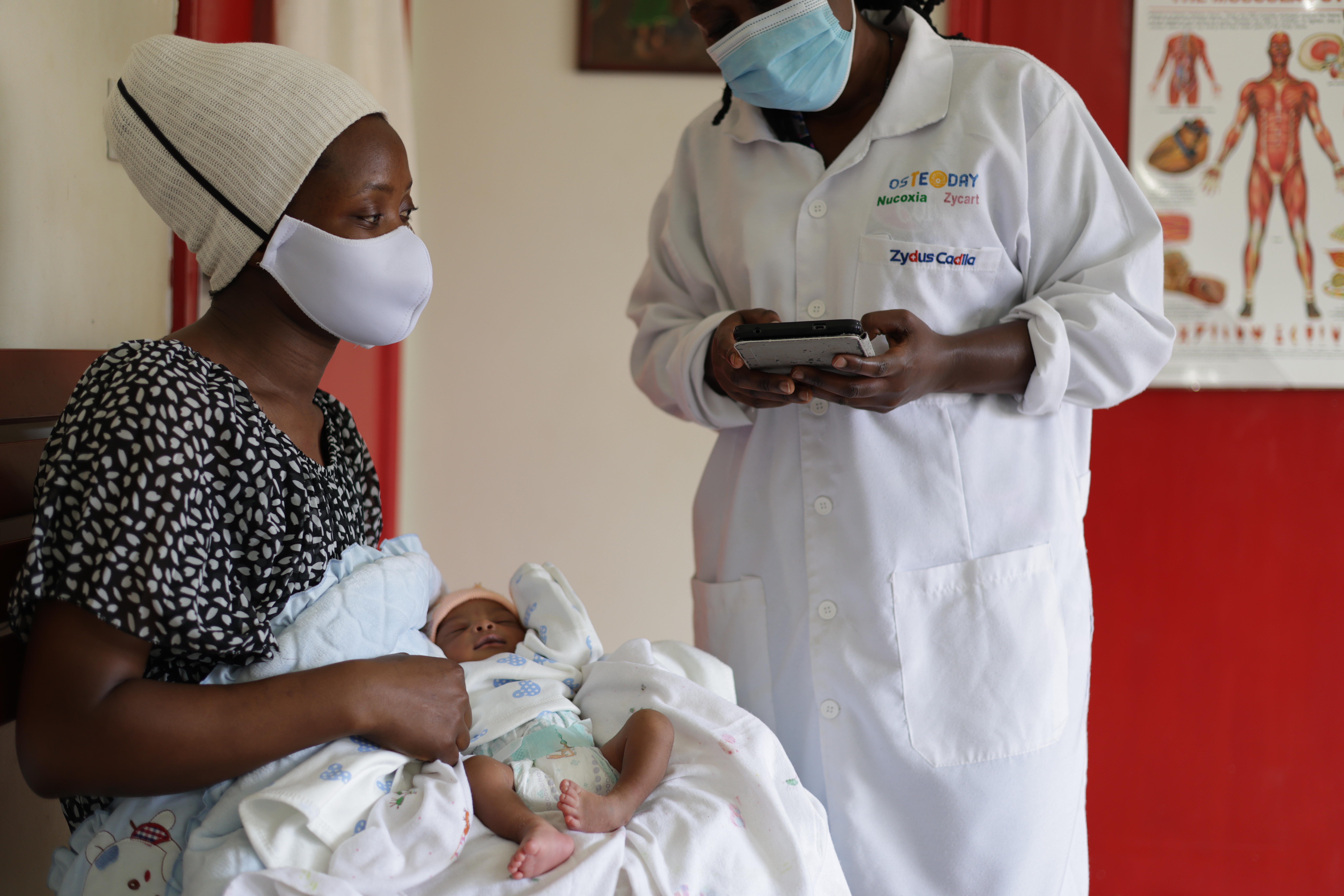 A woman wearing mask holding a newborn baby with clubfoot condition. A healthcare professional is standing next to her speaking.