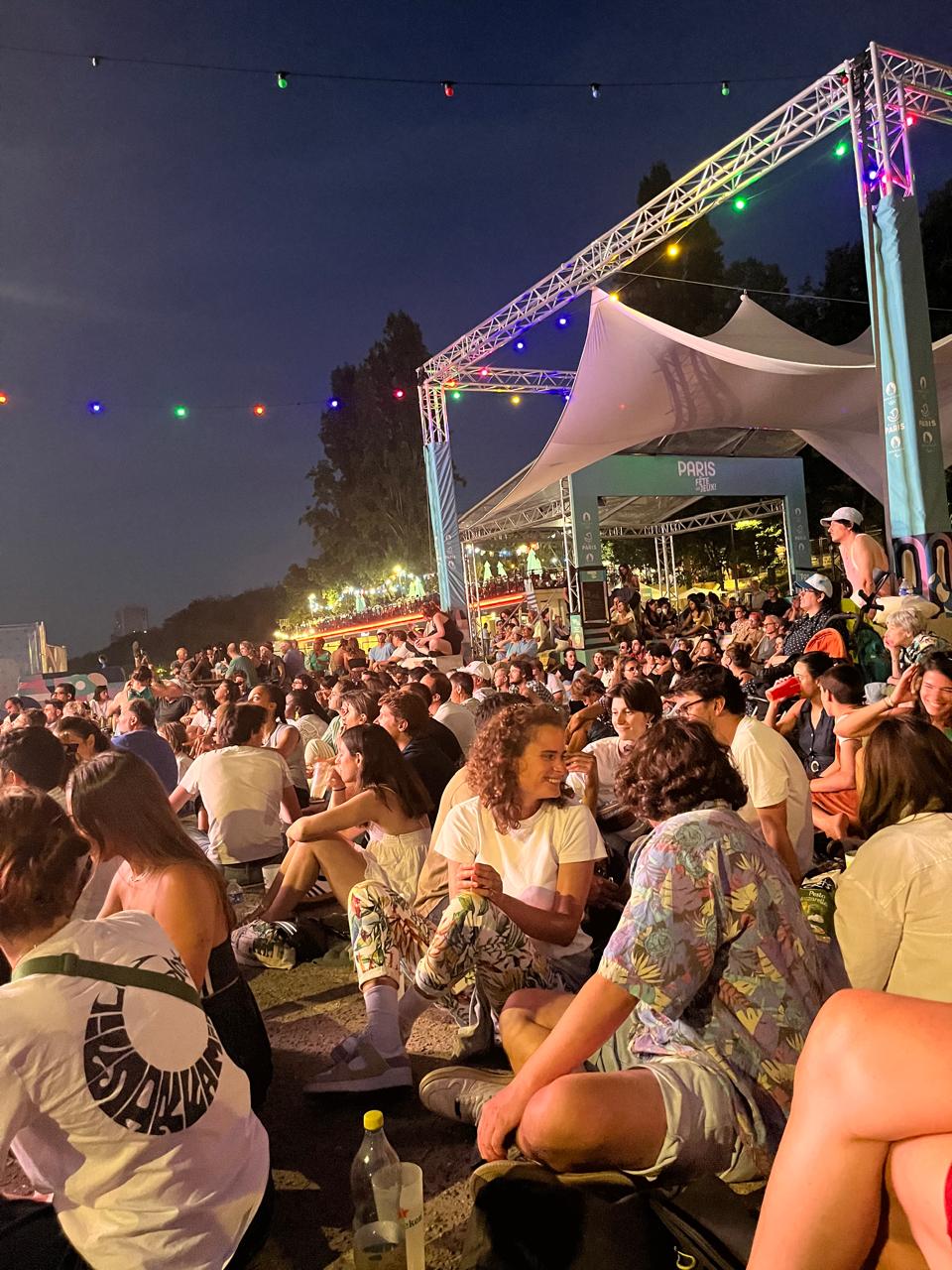 Image of the fan park for the opening ceremony in Paris with large community attendance, sitting watching big screen