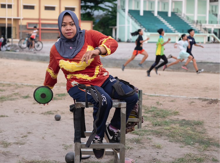 A woman is sat in a sports field in a low resource setting. She appears to be waiting to catch a ball. 