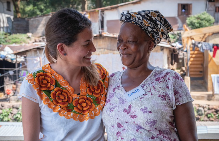 Two woman stand arm in arm smiling towards each other