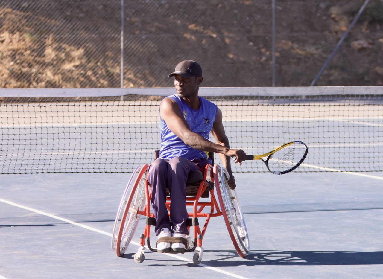 A male wheelchair tennis player wearing a blue sleeveless jersey and black cap prepares to hit a shot on an outdoor tennis court.