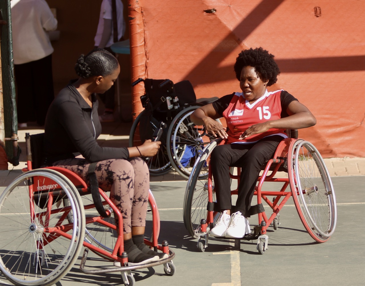 Two women in wheelchairs have a discussion outdoors; one wears a red jersey numbered 15, and the other wears black sportswear.