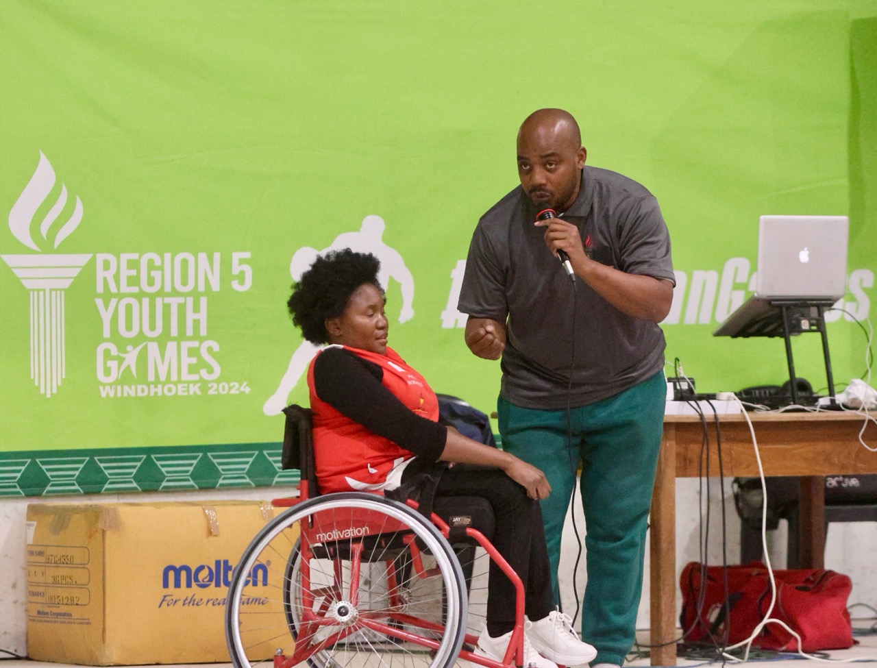 A man speaks into a microphone beside a woman in a wheelchair wearing a red sports jersey, during the Region 5 Youth Games in Windhoek 2025.