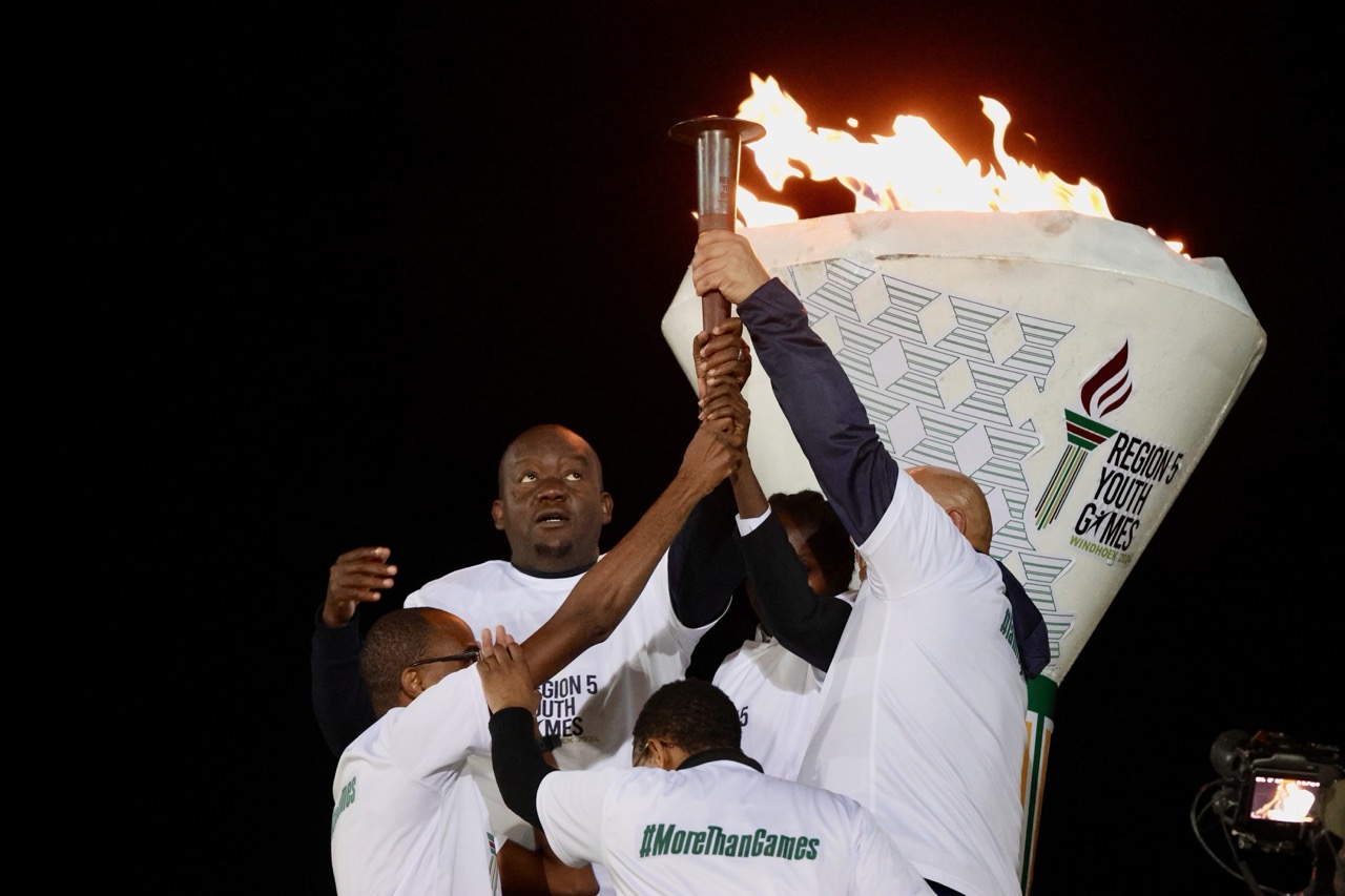 A group of men lift a flaming torch together at the Region 5 Youth Games, symbolizing the opening ceremony.