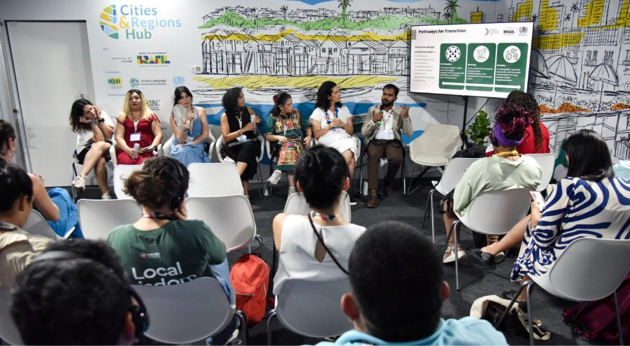 Panel discussion at the Cities & Regions Hub, COP-30, with speakers seated in a semicircle addressing an audience while a slide on &ldquo;pathways for transition&rdquo; displays three green diagrams.