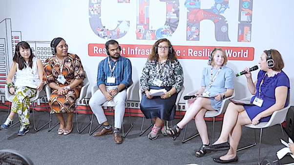 Six panelists sit in a row on white chairs; a woman on the far right speaks into a microphone while others listen with headsets and hold notes. A large backdrop reads &ldquo;CDRI &mdash; Resilient Infrastructure: Resilient World,&rdquo; with stylized city graphics.