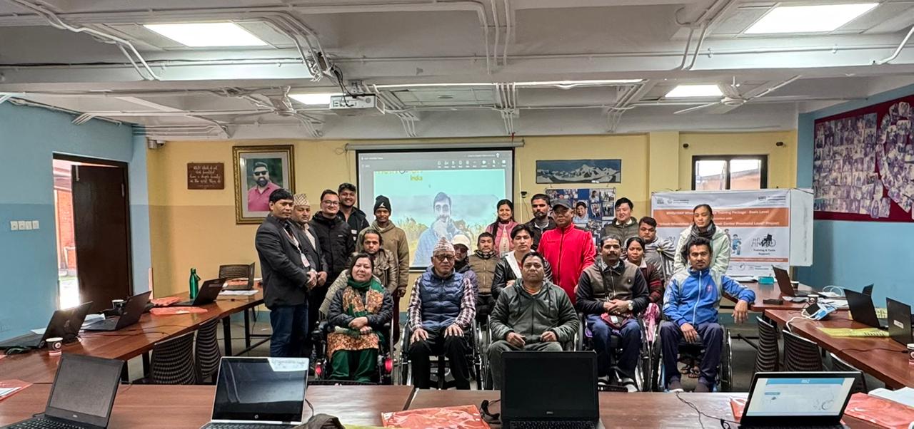  group of people, including several using wheelchairs, pose for a group photo in a classroom-style training room with laptops on tables and a presentation projected on the screen behind them.