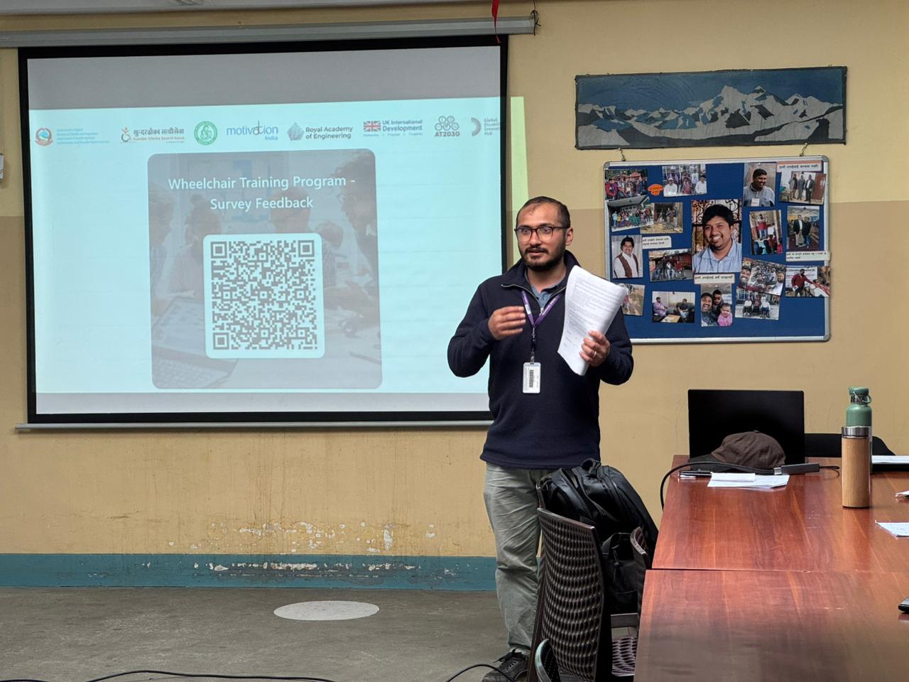 Ram stands presenting in front of a projected slide titled &ldquo;Wheelchair Training Program &ndash; Survey Feedback,&rdquo; holding papers.