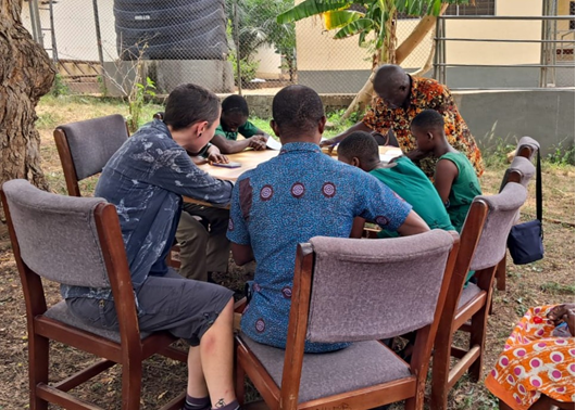 A group of adults and young people with visual impairments sitting around a table. They are sitting outside under a tree. The young people taking part in the most significant change game