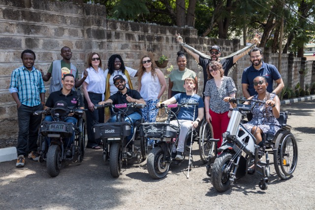 A group of people pose outdoors on a paved street beside a stone wall, with several individuals seated on electric wheelchairs and handcycles labeled “MATT,” while others stand behind them. Trees line the background.
