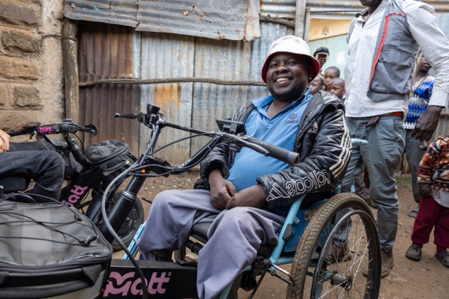 A person sits on a hand‑powered wheelchair next to an electric handcycle labeled “MATT,” on a narrow street with corrugated metal walls and several people standing in the background.