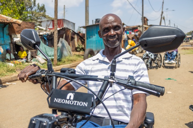 A person sits on an electric handcycle labeled “NEOMOTION” on a dirt road, with multiple handcycles lined up behind them and small roadside structures in the background.