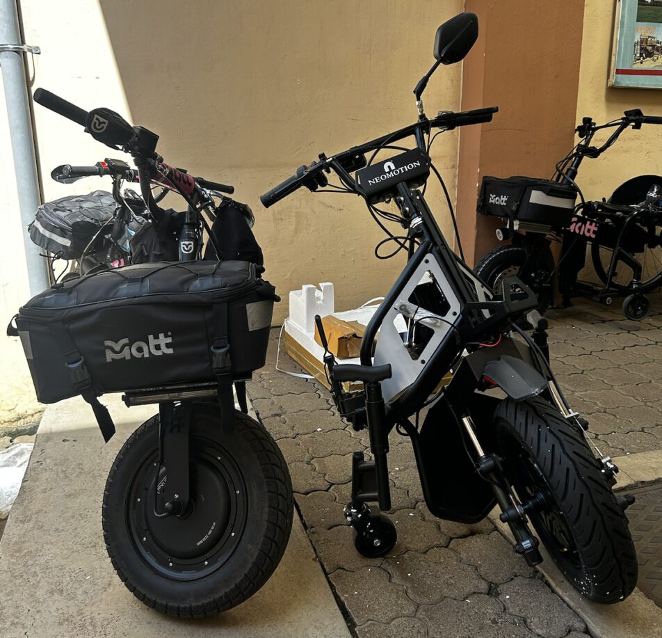 Powered wheelchair attachments with large front wheels and handlebars, branded &ldquo;NeoMotion&rdquo; and &ldquo;Matt,&rdquo; parked against a wall.
