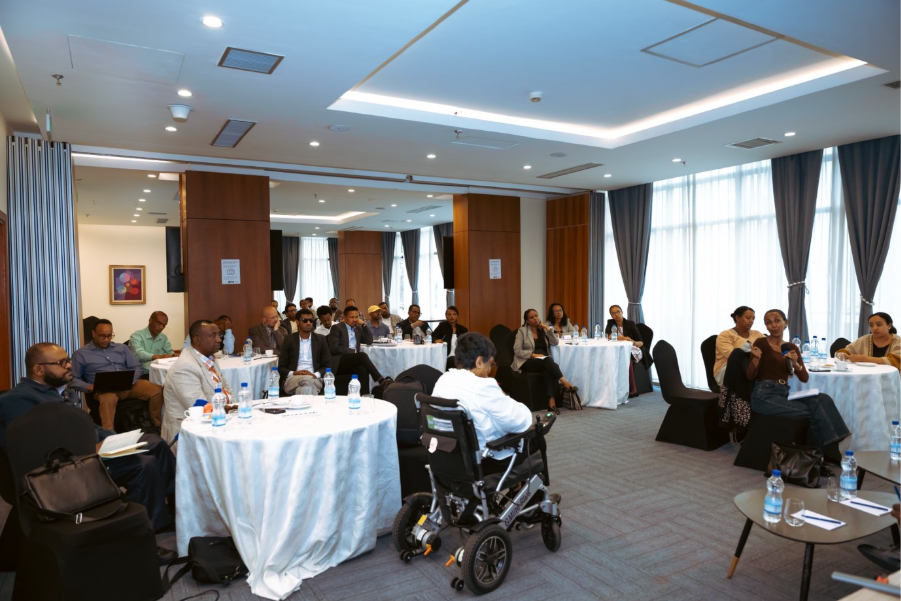A view of the room for the netowkring event, about 25 stakeholders sit around tables, listening to a commentary from one of the event participants. 