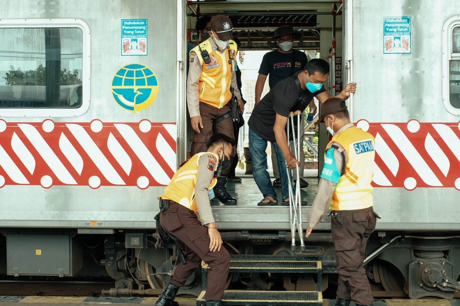 A man using crutches is helped down from a high train to the platform by 3 workers