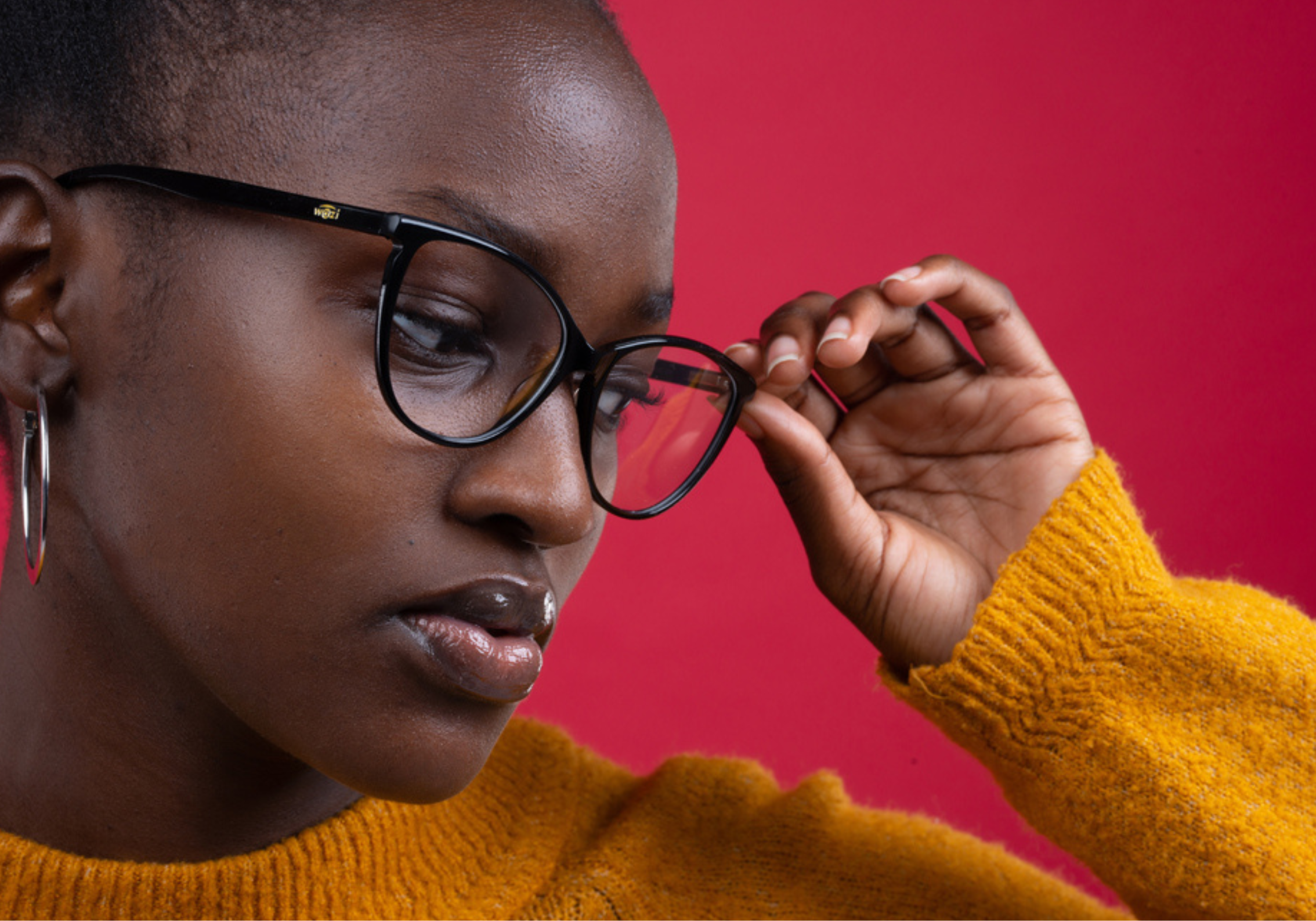 Professonial photograph of a woman posing with stylish glasses. The female in the photo is black and is wearing a bright yellow jumper against a red background