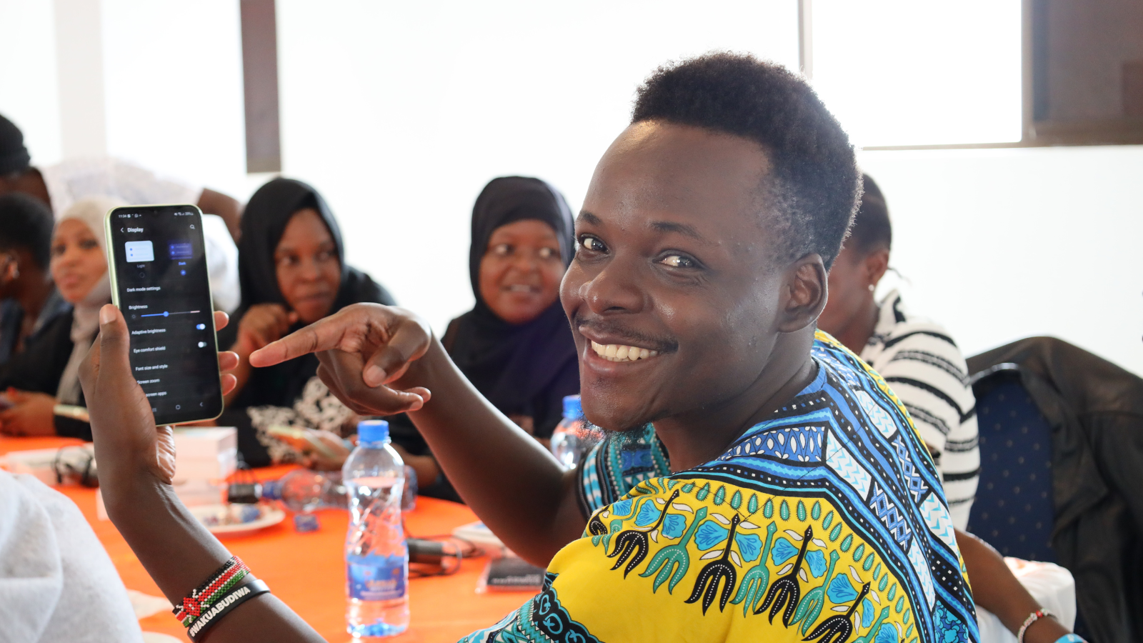 Image in Kenya of a trainer showcasing accessible features on a mobile. Male training is smiling warmly to the camera, in a bright printed top - and pointing to mobile phone.