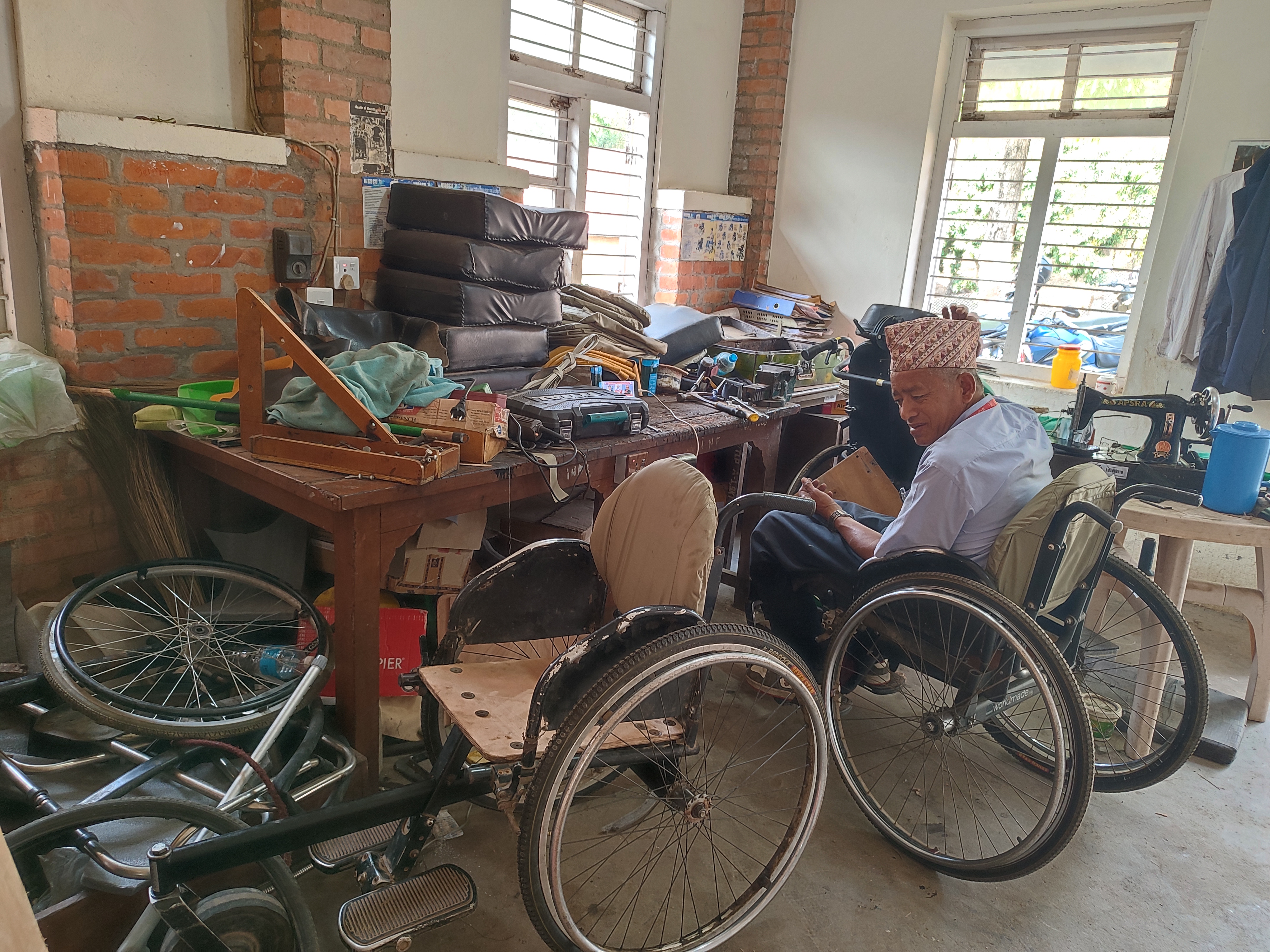 A man in a wheelchair is looking down at a small object in his hands. He sits between a wall with large windows and a cluttered workshop bench. The bench holds various tools, including a drill, a soldering iron, and several stacks of what appear to be seat cushions. The scene suggests a busy wheelchair repair shop. Cover Image